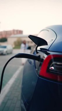 Genç bir kadın bir süpermarketin yakınındaki şarj istasyonunda bir arabayı şarj ediyor. Elektrikli bir arabayı şarj etmek. Elektrikli araba konsepti. Dikey video