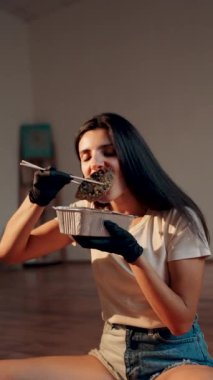 Lotus pozisyonunda otururken yemek çubuklarıyla suşi burger yiyen genç bir kadın. Japon yemeği konsepti. Ellerinde suşi burgeri. Dikey Vedeo 4K.