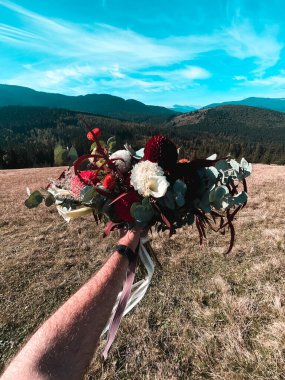 A man's hand holds a craft bouquet of flowers against the background of the mountains. The concept of travel in the mountains clipart