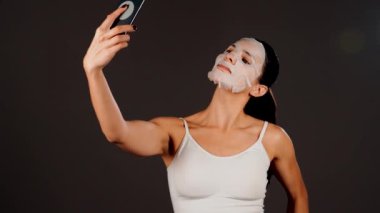 Genç bir kadın koyu bir arka plana kozmetik maske takıyor. Ev yapımı yüz bakımı konsepti. Yüz bakımı için kozmetik maske 4k