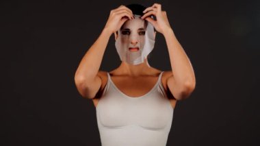Genç bir kadın koyu bir arka plana kozmetik maske takıyor. Ev yapımı yüz bakımı konsepti. Yüz bakımı için kozmetik maske 4k