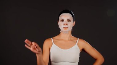 Genç bir kadın koyu bir arka plana kozmetik maske takıyor. Ev yapımı yüz bakımı konsepti. Yüz bakımı için kozmetik maske 4k