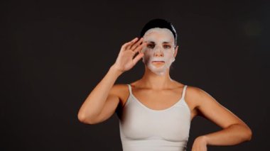 Genç bir kadın koyu bir arka plana kozmetik maske takıyor. Ev yapımı yüz bakımı konsepti. Yüz bakımı için kozmetik maske 4k