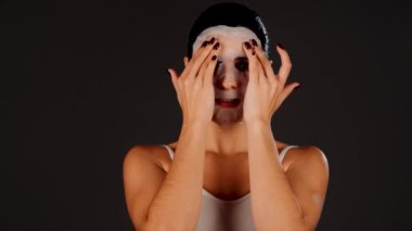 Genç bir kadın koyu bir arka plana kozmetik maske takıyor. Ev yapımı yüz bakımı konsepti. Yüz bakımı için kozmetik maske 4k