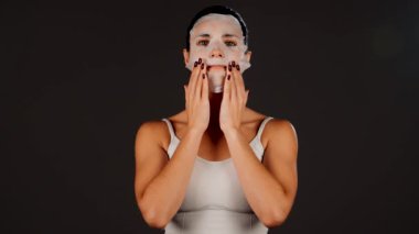 Genç bir kadın koyu bir arka plana kozmetik maske takıyor. Ev yapımı yüz bakımı konsepti. Yüz bakımı için kozmetik maske 4k
