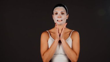 Genç bir kadın koyu bir arka plana kozmetik maske takıyor. Ev yapımı yüz bakımı konsepti. Yüz bakımı için kozmetik maske 4k