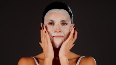 Genç bir kadın koyu bir arka plana kozmetik maske takıyor. Ev yapımı yüz bakımı konsepti. Yüz bakımı için kozmetik maske 4k