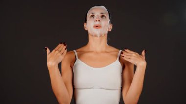 Genç bir kadın koyu bir arka plana kozmetik maske takıyor. Ev yapımı yüz bakımı konsepti. Yüz bakımı için kozmetik maske 4k