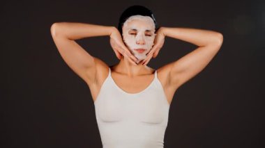 Genç bir kadın koyu bir arka plana kozmetik maske takıyor. Ev yapımı yüz bakımı konsepti. Yüz bakımı için kozmetik maske 4k
