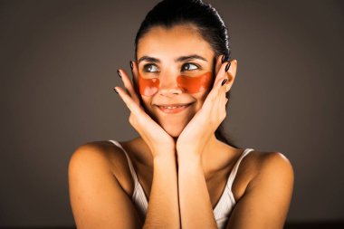 Mükemmel parıldayan derisi ve kameraya poz veren doğal makyajlı genç bir kadın. Gözlerinin altında yorgunluk ve şişkinliği azaltmak için kolajen bantları var. Cilt bakımı. Modern güzellik aletleri konsepti.