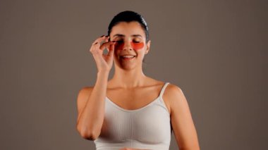 Mükemmel parıldayan derisi ve kameraya poz veren doğal makyajlı genç bir kadın. Gözlerinin altında yorgunluk ve şişkinliği azaltmak için kolajen bantları var. Deri bakımı. Modern güzellik aletleri konsepti 4k.