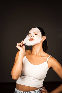 Genç bir kadın koyu bir arka plana kozmetik maske takıyor. Ev yapımı yüz bakımı konsepti. Yüz bakımı için kozmetik maske 4k