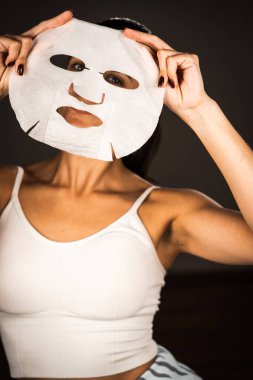 Genç bir kadın koyu bir arka plana kozmetik maske takıyor. Ev yapımı yüz bakımı konsepti. Yüz bakımı için kozmetik maske 4k