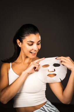 Genç bir kadın koyu bir arka plana kozmetik maske takıyor. Ev yapımı yüz bakımı konsepti. Yüz bakımı için kozmetik maske 4k