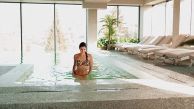 Genç bir kadın dağ manzaralı bir havuzda yüzüyor. Bir kadın kaplıcada dinlenmekten hoşlanır. Otel tatili konsepti 4k