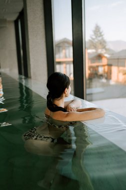 Genç bir kadın, dağları gören kapalı bir havuzda yüzüyor. Bir kadın kaplıcada dinlenmekten hoşlanır. Havuz kenarında dinleniyorum. Otel tatili konsepti 4k