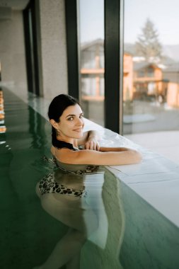 Genç bir kadın, dağları gören kapalı bir havuzda yüzüyor. Bir kadın kaplıcada dinlenmekten hoşlanır. Havuz kenarında dinleniyorum. Otel tatili konsepti 4k