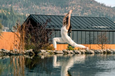 Beyaz takım elbiseli genç bir kadın dağdaki bir evin yakınında yoga yapıyor. Dağlarda meditasyon yapan bir kadın. Yoga ve meditasyon kavramı. Asanas