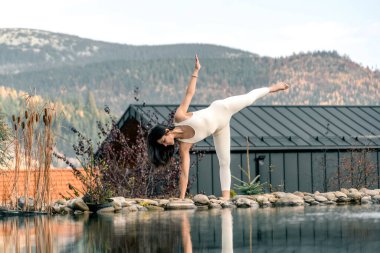 Beyaz takım elbiseli genç bir kadın dağdaki bir evin yakınında yoga yapıyor. Dağlarda meditasyon yapan bir kadın. Yoga ve meditasyon kavramı. Asanas