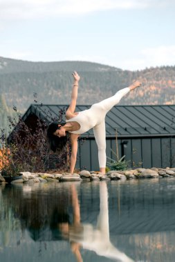 Beyaz takım elbiseli genç bir kadın dağdaki bir evin yakınında yoga yapıyor. Dağlarda meditasyon yapan bir kadın. Yoga ve meditasyon kavramı. Asanas