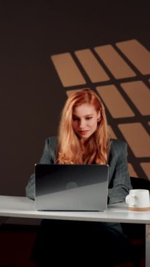 Bir kadın ofisteki başarısına sevinir. Genç bir kadın dizüstü bilgisayarın başında otururken güler ve sevinir. Mutlu bir serbest çalışanın portresi. Çevrimiçi iş kavramı. Dikey video