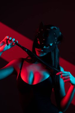 Kedi maskeli bir kadın siyah arka planda kırbaçla poz veriyor. Kamçı bir kadının elindedir. Rol yapma oyunları. BDSM konsepti