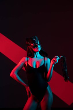 Kedi maskeli bir kadın siyah arka planda kırbaçla poz veriyor. Kamçı bir kadının elindedir. Rol yapma oyunları. BDSM konsepti