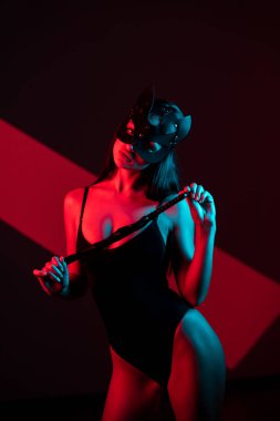 Kedi maskeli bir kadın siyah arka planda kırbaçla poz veriyor. Kamçı bir kadının elindedir. Rol yapma oyunları. BDSM konsepti