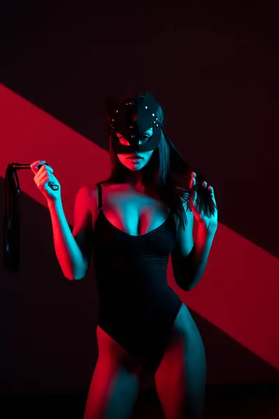 Kedi maskeli bir kadın siyah arka planda kırbaçla poz veriyor. Kamçı bir kadının elindedir. Rol yapma oyunları. BDSM konsepti