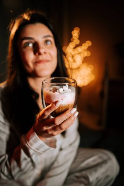 Sıcak kapuçinonun tadını çıkaran bir kadın Marshmallowlu Yatakta dinlenirken Gece Noel süsleriyle çevrili