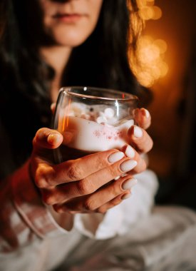 Sıcak kapuçinonun tadını çıkaran bir kadın Marshmallowlu Yatakta dinlenirken Gece Noel süsleriyle çevrili