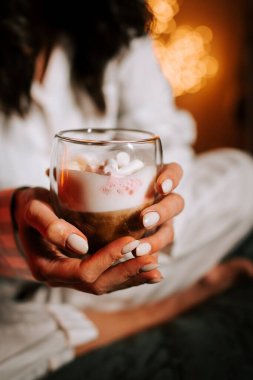 Sıcak kapuçinonun tadını çıkaran bir kadın Marshmallowlu Yatakta dinlenirken Gece Noel süsleriyle çevrili