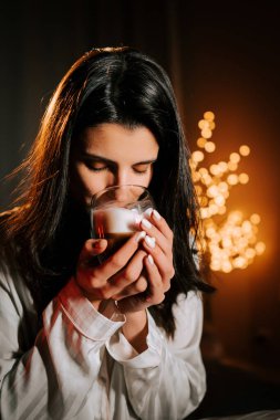 Sıcak kapuçinonun tadını çıkaran bir kadın Marshmallowlu Yatakta dinlenirken Gece Noel süsleriyle çevrili