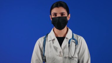 Mavi arka planda siyah bir tıbbi maske takan kadın sağlık çalışanı. Bir doktorun portresi. Viroloji ve salgın konsepti