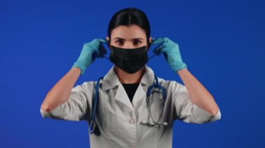 Mavi arka planda siyah bir tıbbi maske takan kadın sağlık çalışanı. Bir doktorun portresi. Viroloji ve salgın konsepti