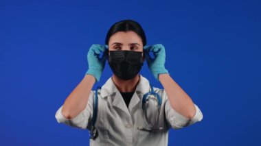 Mavi arka planda siyah bir tıbbi maske takan kadın sağlık çalışanı. Bir doktorun portresi. Viroloji ve salgın konsepti