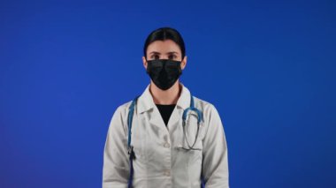 Mavi arka planda siyah bir tıbbi maske takan kadın sağlık çalışanı. Bir doktorun portresi. Viroloji ve salgın konsepti