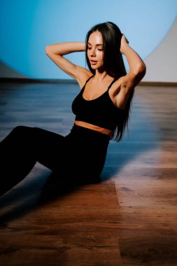 Genç bir kadın atletizm kıyafetleri içinde gün boyunca spor salonunda mavi bir arka planda poz veriyor.
