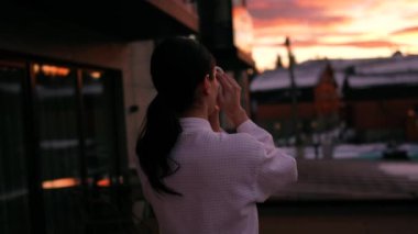 Kadın, sabahlık içinde sabah kahvesinin keyfini çıkarırken huzur içinde güneşin doğuşunu seyrediyor.