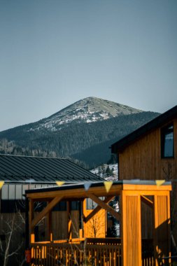 Kış mevsiminde ahşap aksanlı modern mimarinin yer aldığı dağ manzarası, karla kaplı arazi ve ormanları gözler önüne seriyor