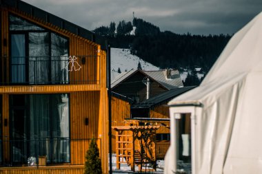 Kış mevsiminde ahşap aksanlı modern mimarinin yer aldığı dağ manzarası, karla kaplı arazi ve ormanları gözler önüne seriyor