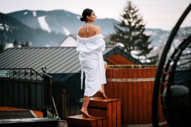 Beyaz mayo giymiş bir kadın, kışın dağlarla çevrili sıcak su fıçısına giriyor..