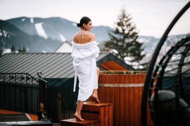 Beyaz mayo giymiş bir kadın, kışın dağlarla çevrili sıcak su fıçısına giriyor..