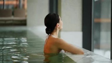 Beyaz mayo giyen kadın kış tatili kaçamağı sırasında karlı dağ manzaralı spa havuzunun keyfini çıkarıyor.