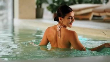 Beyaz mayo giyen kadın kış tatili kaçamağı sırasında karlı dağ manzaralı spa havuzunun keyfini çıkarıyor.