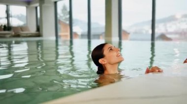 Beyaz mayo giyen kadın kış tatili kaçamağı sırasında karlı dağ manzaralı spa havuzunun keyfini çıkarıyor.