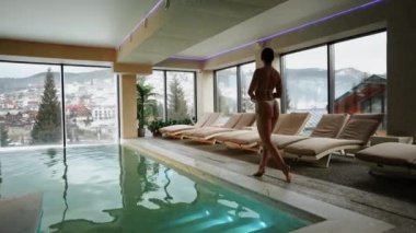 Beyaz mayo giyen kadın kış tatili kaçamağı sırasında karlı dağ manzaralı spa havuzunun keyfini çıkarıyor.