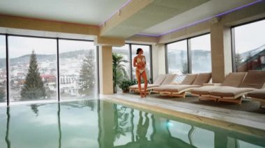 Beyaz mayo giyen kadın kış tatili kaçamağı sırasında karlı dağ manzaralı spa havuzunun keyfini çıkarıyor.
