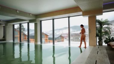 Beyaz mayo giyen kadın kış tatili kaçamağı sırasında karlı dağ manzaralı spa havuzunun keyfini çıkarıyor.