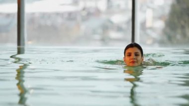 Beyaz mayo giyen kadın kış tatili kaçamağı sırasında karlı dağ manzaralı spa havuzunun keyfini çıkarıyor.